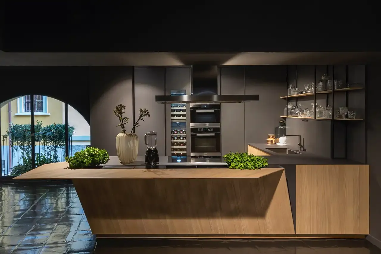 A light oak kitchen island with an angled front, set against dark grey cabinets with built-in ovens and open shelving